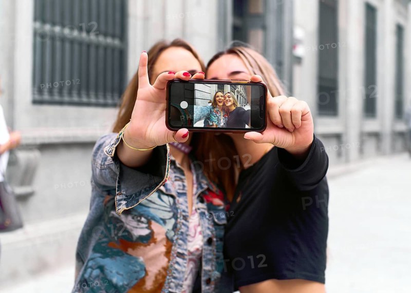 Two young friends capture a joyful selfie with a smartphone on a city street. The phone screen reveals their smiling faces, symbolizing friendship and connection