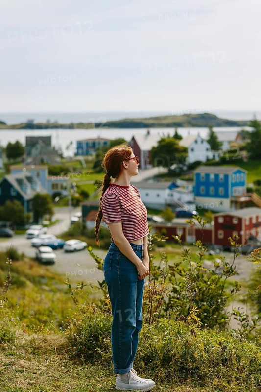 Side view of a red hair woman, captured from the side with a peaceful stance, gazes over the charming coastal town of Trinity, Newfoundland & Labrador, showcasing the scenic beauty and tranquil surroundings