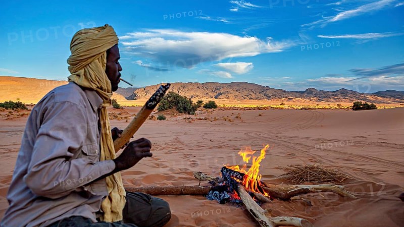 A man sits around a campfire in a vast desert landscape under a clear sky, Tuareg around a campfire smoking a cigarette in the Sahara desert in Algeria