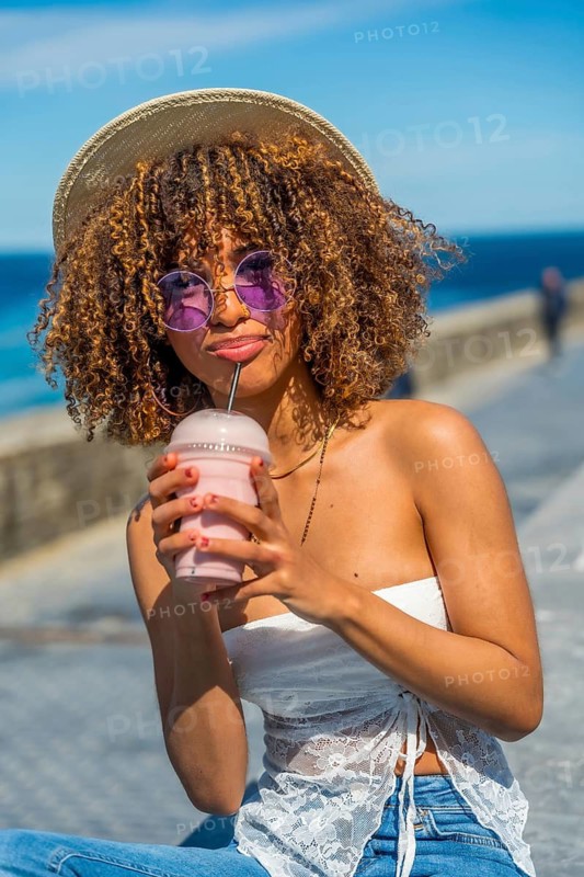 Vertical portrait of a young stylish latin woman drinking iced juice sitting next to the sea