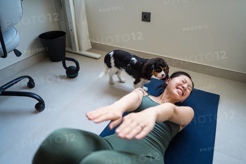 A young Asian woman attempts her home workout, lying on a yoga mat, as her playful dog interrupts her exercise session, creating a charming and joyful moment of laughter