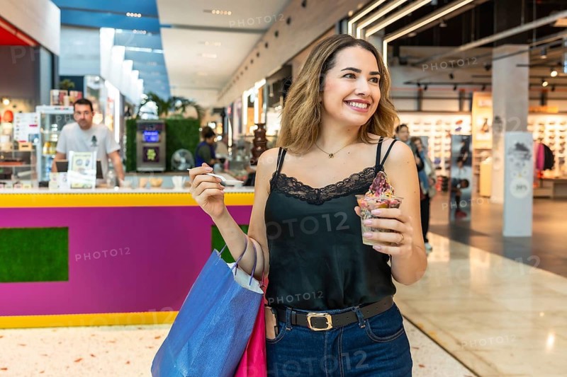 Young woman smiling and walking with shopping bags, eating frozen yogurt dessert in a modern shopping mall environment, enjoying leisure and retail therapy