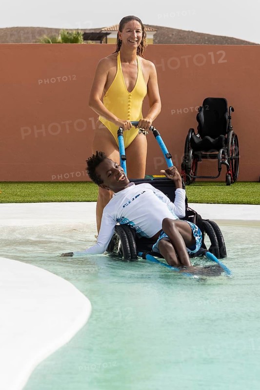 A joyful scene of a woman in a yellow swimsuit pushing a black boy in a specialized wheelchair with cerebral palsy in pool. A standard wheelchair is visible in the background