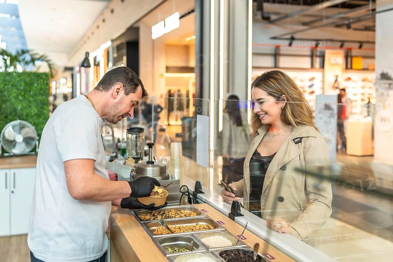 Vendor serving a personalized acai bowl to a smiling female customer at a healthy food kiosk inside an urban shopping mall, offering fresh and nutritious options