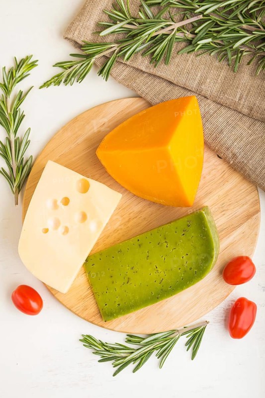Green basil cheese and various types of cheese with rosemary and tomatoes on wooden board on a white wooden background. Top view, copy space, selective focus