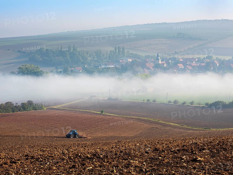 View of the village of Rhoden in autumn, fog sweeping through the valley, a tractor ploughs a field, Osterwieck, Harz, Harz foothills, Saxony-Anhalt, Germany