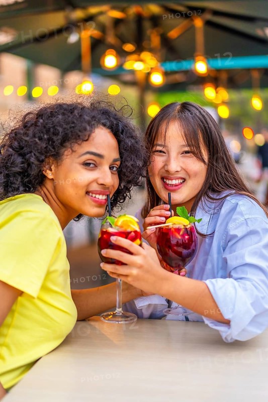 Vertical portrait of lesbian multi-ethnic couple enjoying cocktails smiling at camera in an outdoor terrace