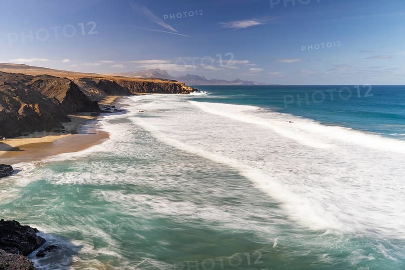 A stunning view of La Pared Beach in Fuerteventura with turquoise ocean waves crashing against the rugged coastline. The vast sky and cliffs highlight the beauty of this untouched paradise