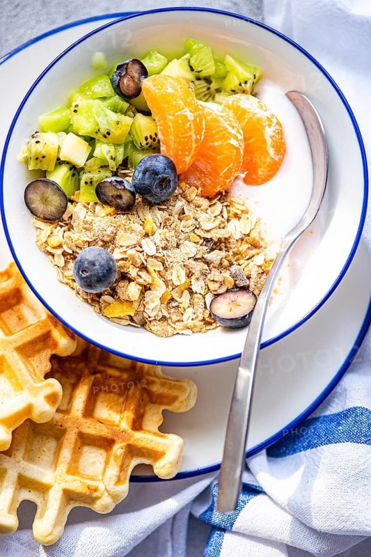 A vibrant breakfast bowl filled with kiwi, mandarin, and blueberries atop yogurt and granola, accompanied by waffles. Perfect for a healthy start to the day