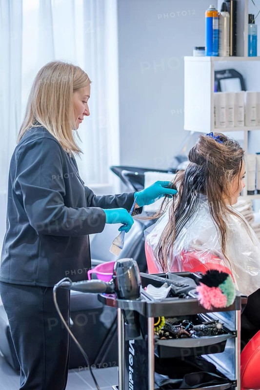 A woman receives a keratin hair treatment at a beauty salon. The hair stylist works carefully, using tools and techniques for hair straightening. The salon atmosphere is busy yet focused on hair care