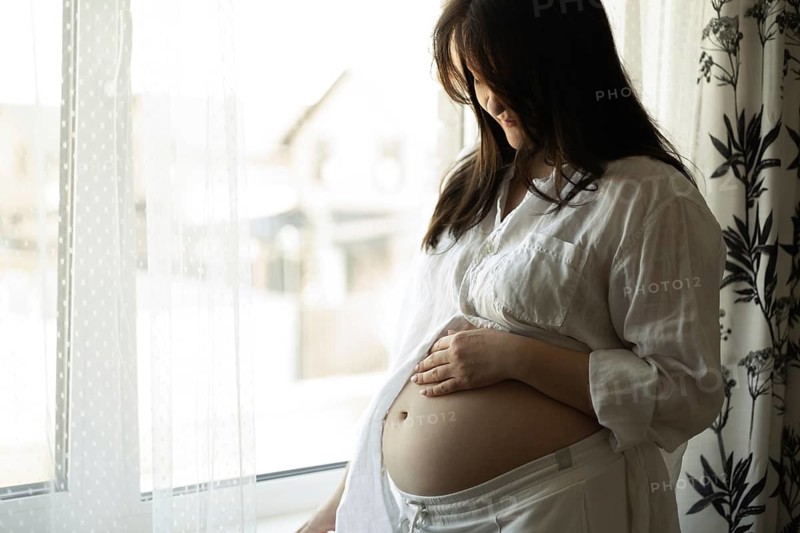 A serene pregnant woman stands by a sunlit window, gently holding her belly. She is immersed in thoughts of motherhood and the exciting journey that lies ahead