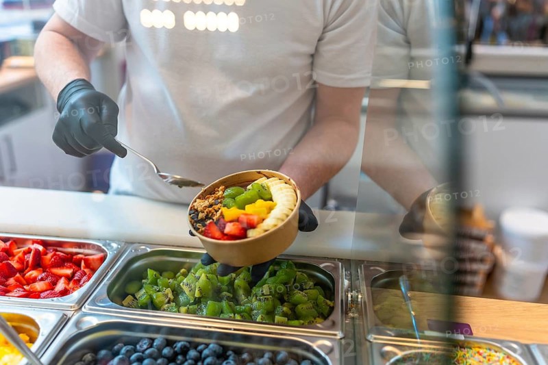 Person wearing black gloves holding a kraft paper bowl filled with acai, fresh strawberries, kiwi, mango, banana slices, and granola, preparing a healthy superfood snack or breakfast