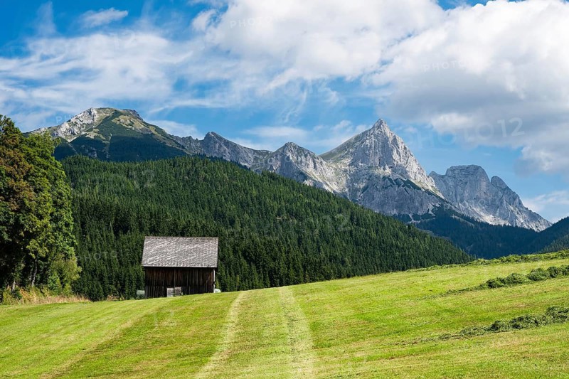 A picturesque landscape of the Austrian Alps featuring traditional wooden barns and houses nestled in lush green meadows beneath jagged, towering mountain peaks in Austria