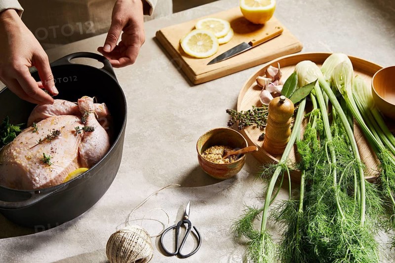 A cook seasons a whole chicken with fresh herbs and lemon in a kitchen setting. Fennel, garlic, and other seasonings are prepared on a table for a flavorful meal