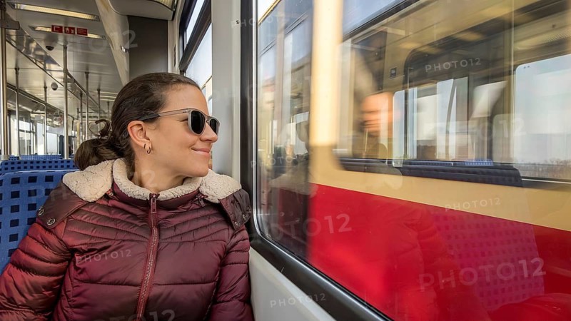 A woman in sunglasses and a cozy jacket enjoys a scenic train ride on a sunny day, gazing out the window at passing landscapes. The train offers a reflective, serene travel experience