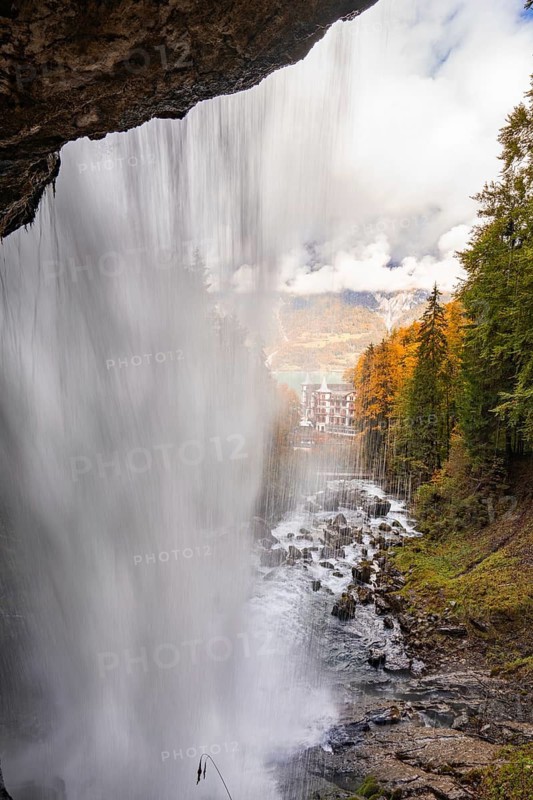 View through a waterfall onto an autumn landscape with mountains and a building, Lake Brienz, Giessbach Waterfall, Switzerland