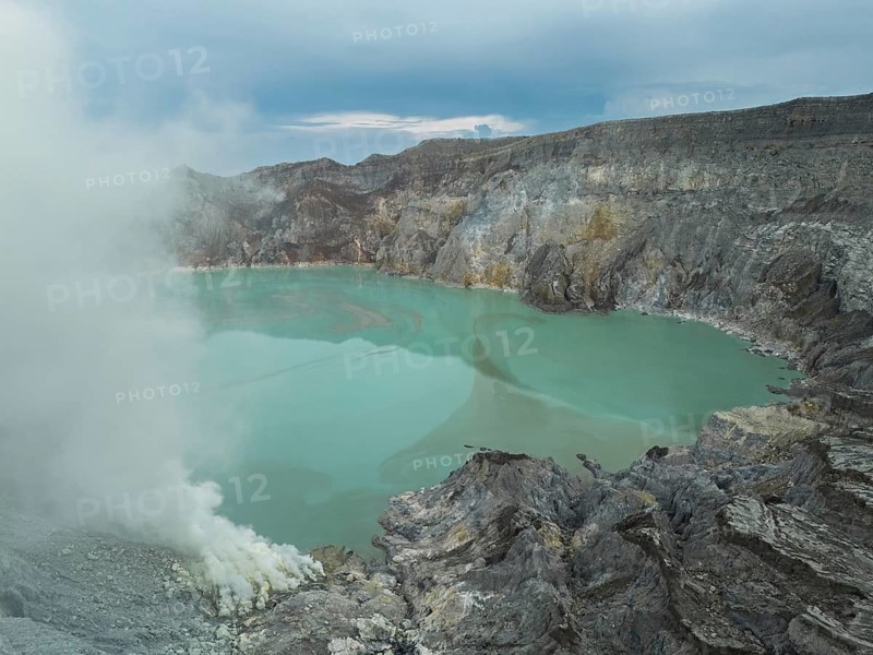 A bright turquoise acidic lake lies inside Ijen volcano, with sulfur gas rising from vents on the rocky edge. A dramatic volcanic scene in East Java, Indonesia