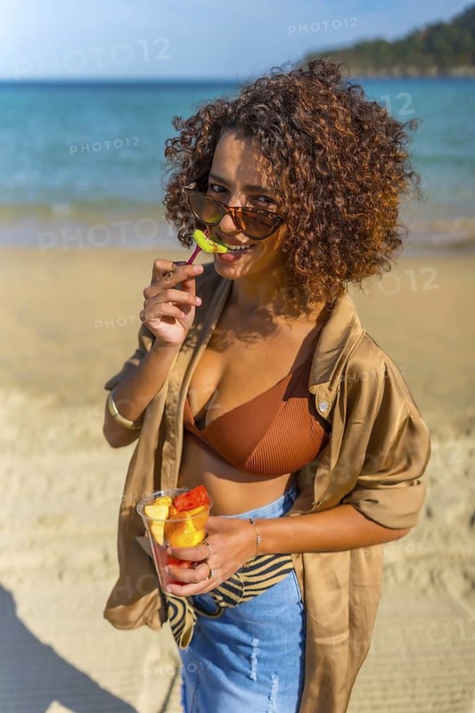 Vertical photo of a Confident arab woman biting a slice of tropical fruit on the beach