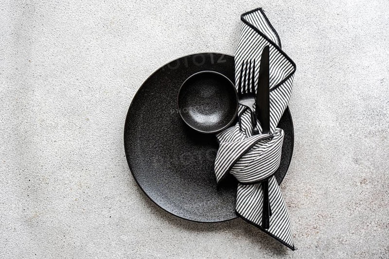 A minimalist black and white table setting features a textured black plate, a striped napkin, and sleek cutlery, creating an elegant dining atmosphere