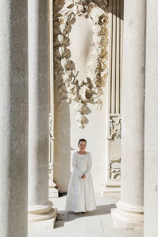 A young girl in a First Communion dress stands between majestic stone columns in a sunlit setting, with intricate architectural designs in the background