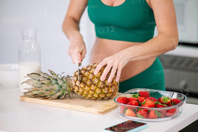 A pregnant woman in a green yoga outfit is cutting a fresh pineapple on a wooden board. Strawberries in a bowl and a jug of milk are on the white kitchen counter