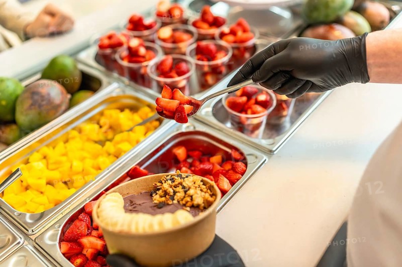 Gloved hand scooping fresh strawberries to top an acai bowl with banana and granola, featuring a selection of diced mango and pre portioned strawberries in a food service setting