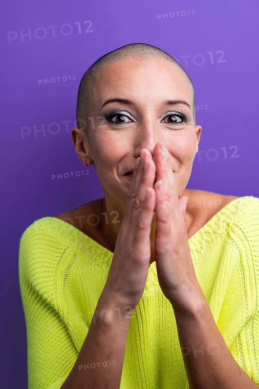 A middle aged woman with a shaved head is laughing and covering her mouth with her hands, dressed in a vibrant yellow sweater against a purple background, emanating happiness and confidence