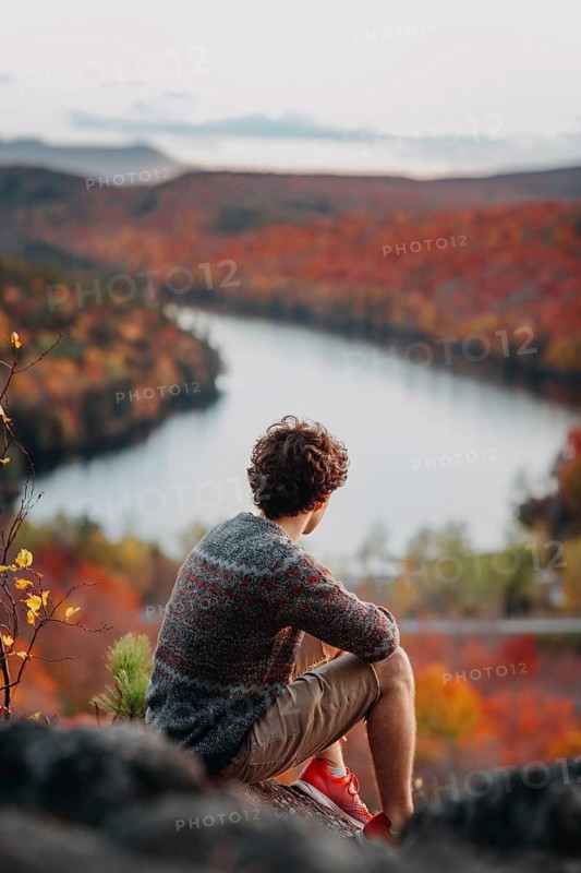 A man sits on a rocky ledge overlooking a river winding through a vibrant, colorful autumn landscape in Quebec, Canada