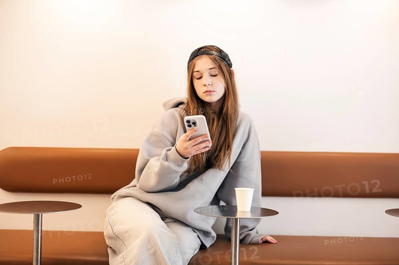 A teenage girl sitting on a stylish bench, wearing casual clothing and a cap, attentively using a smartphone. A white paper cup rests on a nearby table, adding to the laid-back ambiance