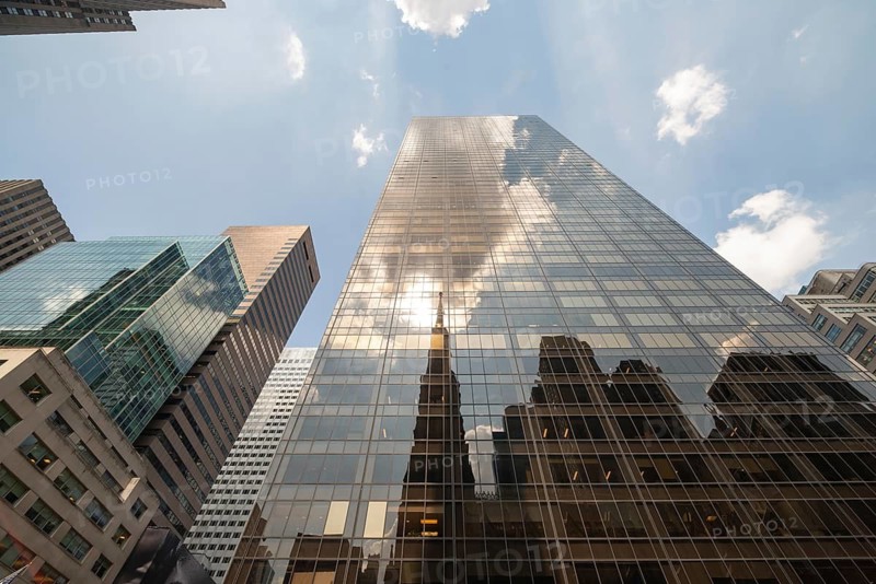 Upward view of a modern glass building in Manhattan, showcasing a reflection of the surrounding skyscrapers against a backdrop of a clear blue sky