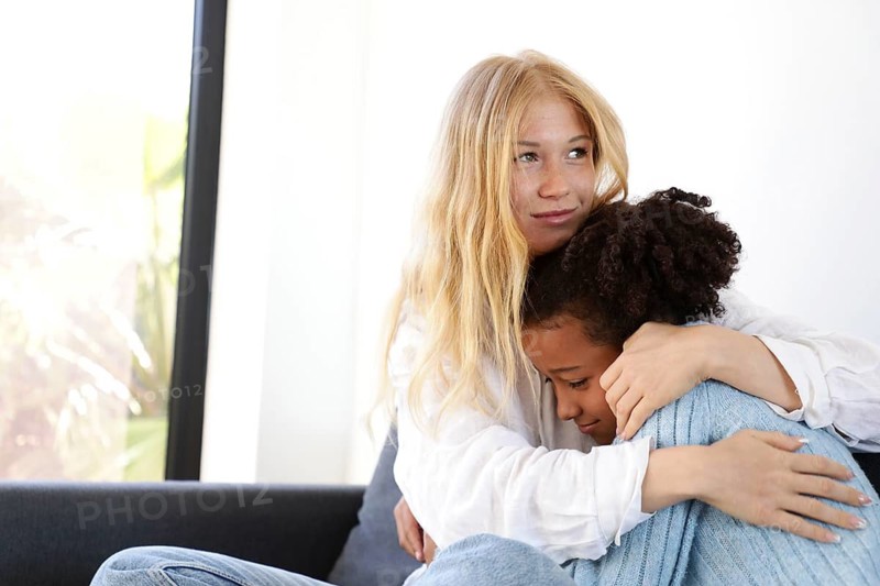 A young woman lovingly embraces a young girl on the couch, offering comfort and reassurance. The warm moment highlights trust, care, and emotional connection in a calm home setting
