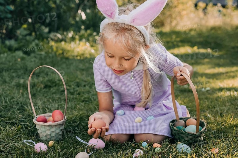 A young girl wearing bunny ears excitedly searches for colorful eggs during an Easter egg hunt, capturing the joy of the festive occasion in a sunlit garden