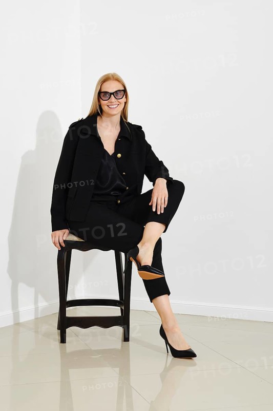 Fashionable beautiful smiling woman with sunglasses in black fashion blazer and trousers sits on wooden chair in bright room