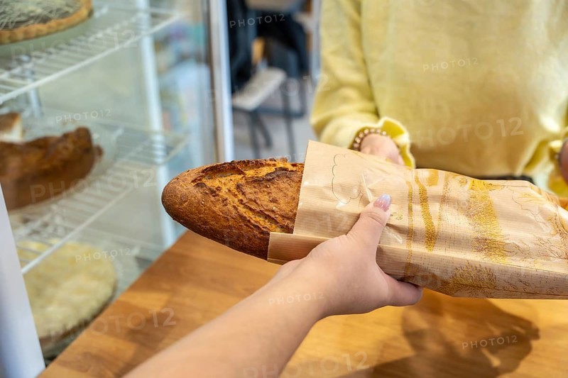 Customer's hand reaching for a warm, crusty baguette wrapped in a brown paper bag, handed over by a bakery employee, highlighting the fresh daily offerings of a local shop