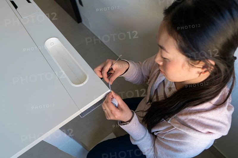 A young Asian woman is intricately involved in a DIY furniture project at home, skillfully assembling a sleek white desk with attention to detail, highlighting the art of craftsmanship