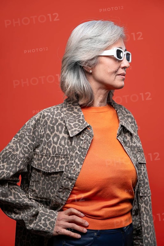 Asian Chinese senior woman exudes confidence with her stylish gray hair, wearing a leopard print jacket and orange sweater, set against a bold red background