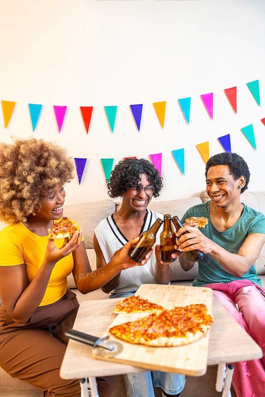 Vertical photo of three diverse male and female friends eating pizza sitting on the sofa together at home