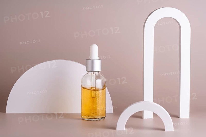 A cosmetic in glass dropper bottle filled with golden oil stands against a beige background, complemented by white geometric shapes, highlighting a minimalist and elegant design