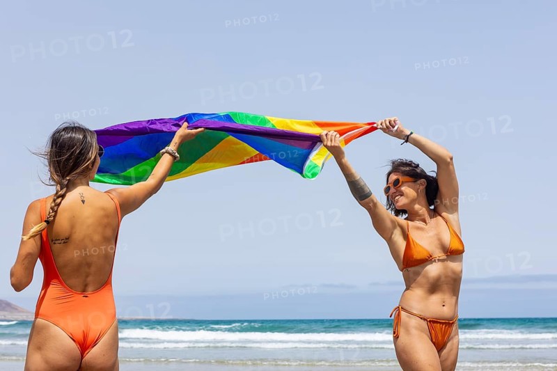 Couple in orange swimwear joyfully wave a rainbow pride flag on a sunny beach. The scene captures freedom, diversity, and summertime happiness