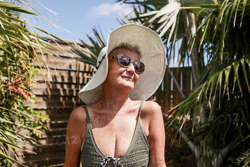A senior woman is pictured in formal summer attire, smiling under her sun hat, surrounded by lush tropical greenery