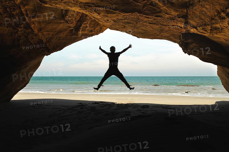 A man jumps inside a cave at Mancora Beach, Peru, creating a striking silhouette against the ocean backdrop. The cave frames the sea and sky, capturing a moment of joy and freedom