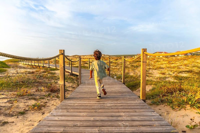 Young boy running on a wooden boardwalk leading through sand dunes towards the beach, experiencing joyful childhood moments during a sunset holiday in seignosse, france