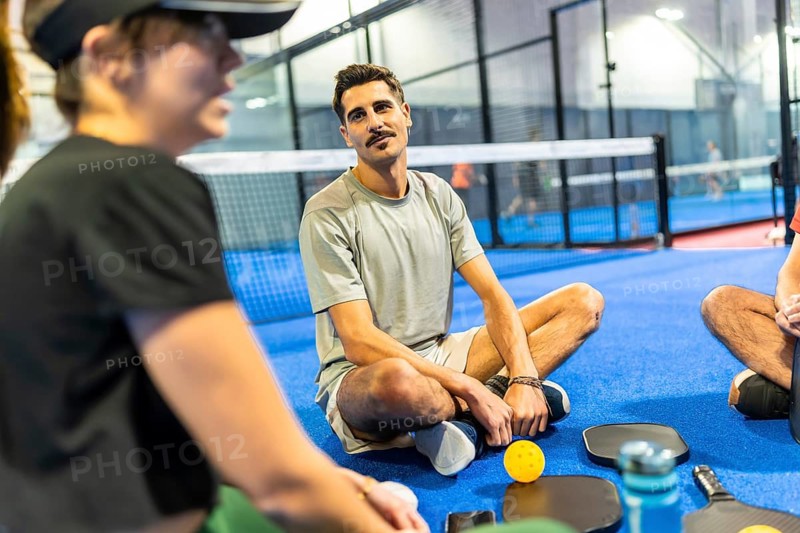 Friends resting and chatting on a blue indoor pickleball court after a game, enjoying conversation, teamwork, fitness and relaxed social bonding during a casual sports break