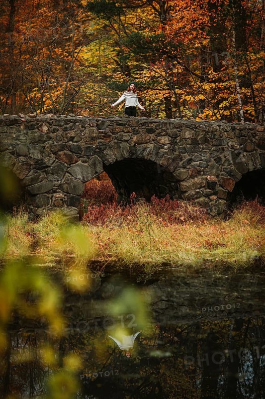 A woman stands serenely on a stone bridge in Quebec, Canada, surrounded by the vibrant colors of autumn. The scene reflects the peacefulness of a quiet fall day