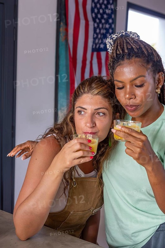 Two women share a warm embrace while enjoying a drink, symbolizing the strength and love within a lesbian family. The American flag in the background reinforces pride and liberty