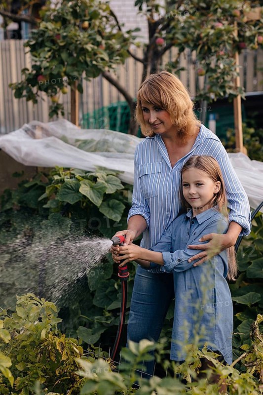 A mother and daughter enjoy a sunny day gardening together. They water plants in their small greenhouse, surrounded by greenery, wearing casual outfits and sharing joyful moments
