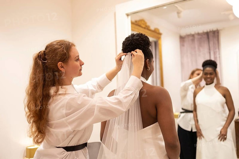 A bride in an elegant gown prepares for her wedding day with the help of a stylist adjusting her veil, reflecting excitement and anticipation