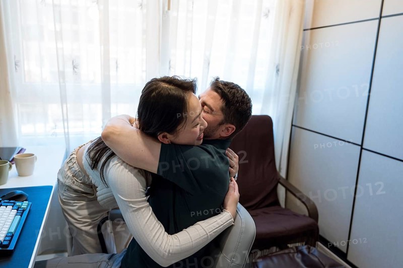 A diverse couple shares a joyful embrace in their cozy home, surrounded by natural light. Their expressions reflect happiness and warmth, creating a moment of genuine connection