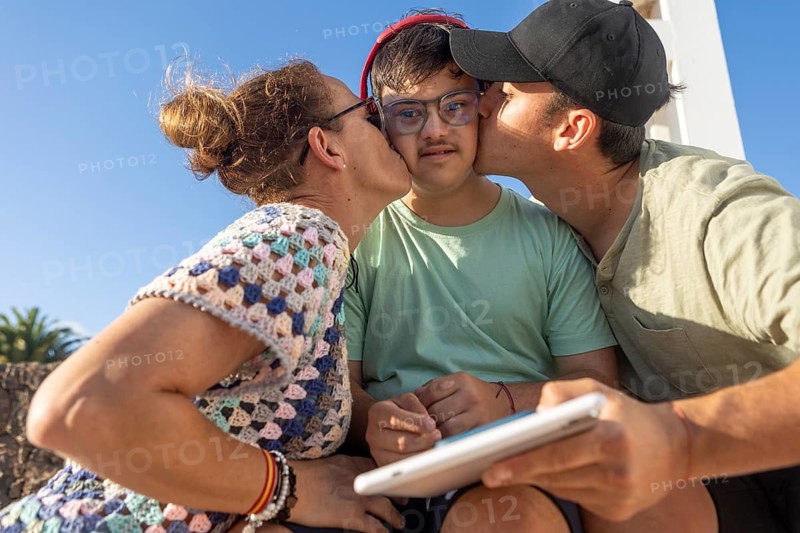 Mother and brother share a sweet moment kissing their Down Syndrome teenager outdoors, creating a warm and loving atmosphere full of affection, connection, and togetherness