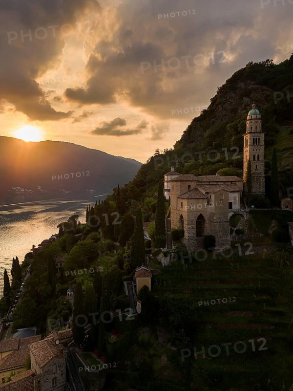A breathtaking sunset over Morcote, Switzerland, capturing the serene view of a historic church perched on a hill overlooking Lake Lugano, with the sun setting behind mountains